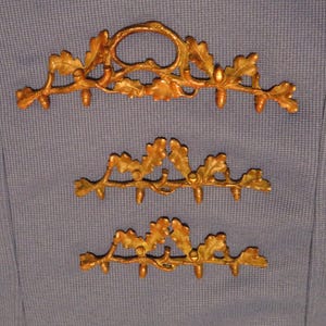 May include: Three decorative golden oak leaf and acorn wall hooks. The top hook has a circular design, while the other two are smaller. The hooks are made of metal and are mounted on a blue fabric background.