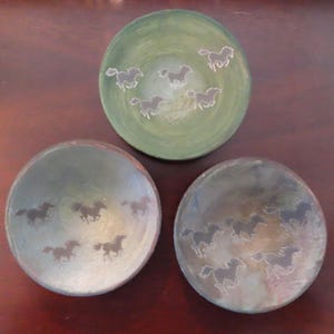 May include: Three decorative ceramic bowls with a horse motif. The bowls are in shades of green and gray, with a running horse design etched into the surface. The bowls are round and have a shallow depth, suitable for serving snacks or displaying as decor.