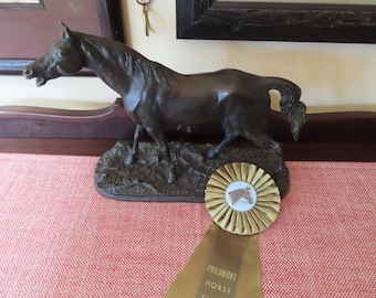 May include: A bronze horse sculpture on a base, with a horse show ribbon in the foreground. The sculpture depicts a horse in motion. The ribbon is gold with a circular center featuring a horse's head.