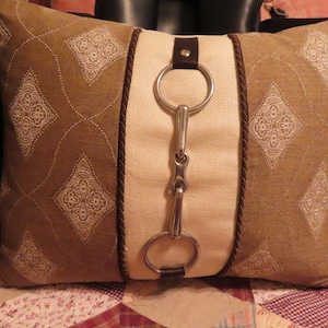 May include: A rectangular throw pillow featuring a central cream-colored vertical panel flanked by two brown sections with a white diamond-shaped pattern. A silver-toned metal horse bit is attached to the center panel with dark brown leather straps.