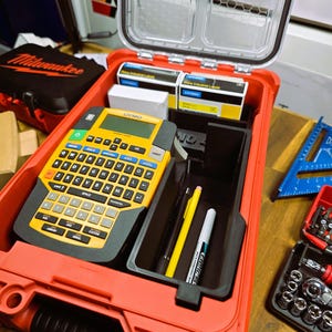 Puede incluir: Una caja de herramientas roja abierta con una rotuladora DYMO amarilla y negra, bolígrafos y cartuchos de etiquetas. Se ve una caja negra de Milwaukee en el fondo.