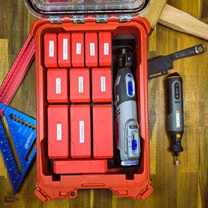 Può includere: Una valigetta portautensili in plastica rossa contenente uno strumento rotativo Dremel e vari accessori per la levigatura e la smerigliatura. La valigetta è etichettata con il contenuto, tra cui "Scultura", "Levigatura 180", "Levigatura 120", "Levigatura 80", "Smerigliatura", "Lucidatura" e "Varie".