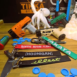 May include: A collection of toy guns in various colours, including orange, green, and tan, alongside keychains with text such as "TAKATA", "REMOVE BEFORE FLIGHT", and "[HOONIGAN]". Also, blue bottle caps and a white octopus lamp.