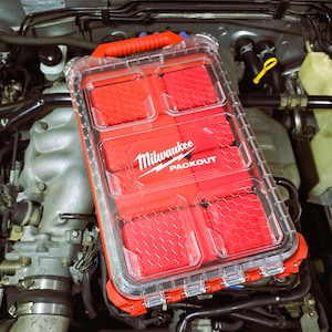 May include: A red and clear Milwaukee Packout storage container with a handle. The container has multiple compartments with a red honeycomb pattern. The Milwaukee logo is visible on the front of the container.
