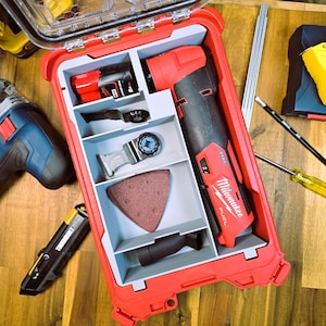 May include: A red Milwaukee FUEL oscillating tool with a battery and accessories is organized in a red and gray tool box. The tool box has a clear lid and is open. The tool is in a compartment with a gray foam insert. The accessories include a sanding pad, a blade, and a battery.