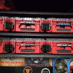 Può includere: Una serie di contenitori in plastica rossa e nera con coperchi trasparenti, organizzati su scaffali. Ogni contenitore presenta un'etichetta che ne identifica il contenuto, tra cui BATTERIES, DREMEL, Detail Sander, M12 Detail Polisher e M12 Multi-Tool, con chiusure nere.
