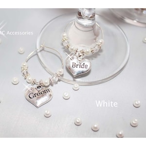 May include: Two silver wine glass charms, one with a heart pendant that says "Bride" and the other with "Groom". Both charms are adorned with pearls and crystals. The background is white with scattered pearls.