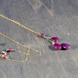 May include: A delicate gold necklace with a chain and a pendant featuring three teardrop-shaped, deep magenta gemstones. The necklace is displayed on a wooden surface, with the text "Plum Blossom Cottage" in the lower left corner.