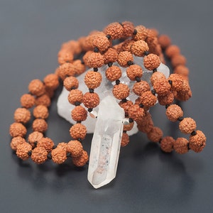 May include: A clear quartz crystal pendant hangs from a brown mala bead necklace. The necklace is made of natural wood beads and is tied with a black cord.