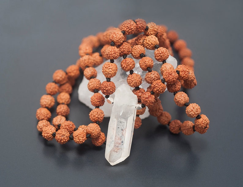 May include: A clear quartz crystal pendant hangs from a brown mala bead necklace. The necklace is made of natural wood beads and is tied with a black cord.