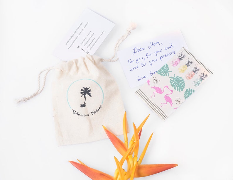 May include: A small white drawstring bag with a palm tree logo and the text "Bahamas Vintage" printed on it. A handwritten note with the text "Dear Mom, For you, for your soul and for your passion Love from" is next to the bag. A bright orange flower is in the bottom right corner of the image.