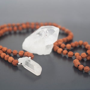 May include: A brown mala bead necklace with a clear quartz crystal pendant. The necklace is made of natural wood beads and has a simple design.