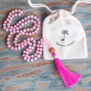 May include: A pink rose quartz mala necklace with a brown wooden bead and a pink tassel. The necklace is in a white drawstring bag with the text "Bohemian Treasure".