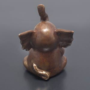 May include: A small, bronze elephant figurine with its trunk raised in the air. The elephant is sitting with its legs tucked underneath its body. The figurine has a detailed, realistic design.