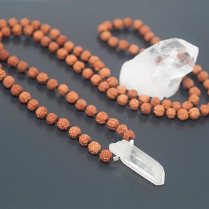 May include: A brown wooden mala bead necklace with a clear quartz crystal pendant. The necklace is laid out on a dark gray surface.