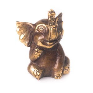 May include: A small, gold-colored elephant figurine with a raised trunk and a smiling face. The figurine is made of metal and has a detailed design.