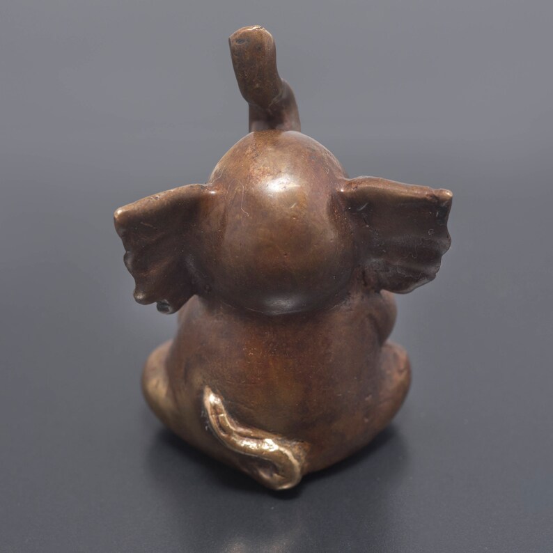 May include: A small, bronze elephant figurine with its trunk raised in the air. The elephant is sitting with its legs tucked underneath its body. The figurine has a detailed, textured surface.