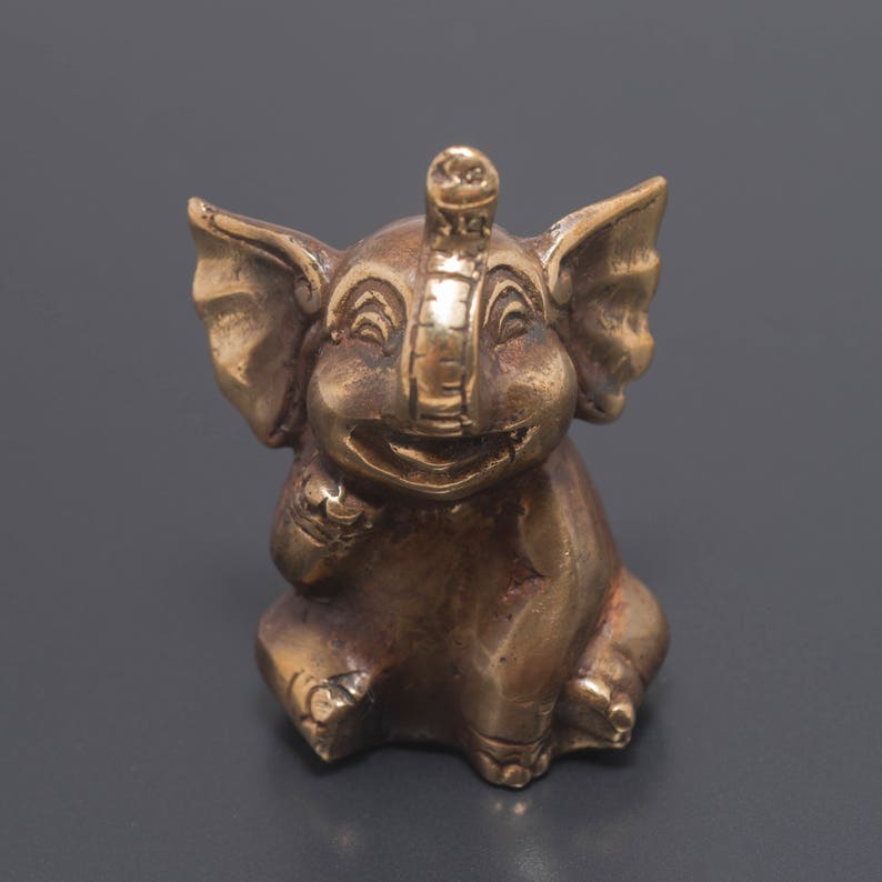 May include: A small, gold-colored metal elephant figurine with a smiling face and raised trunk. The elephant is sitting on its hind legs with its front legs raised.