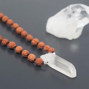 May include: A brown wooden mala bead necklace with a clear quartz crystal pendant. The necklace is lying on a dark gray surface.