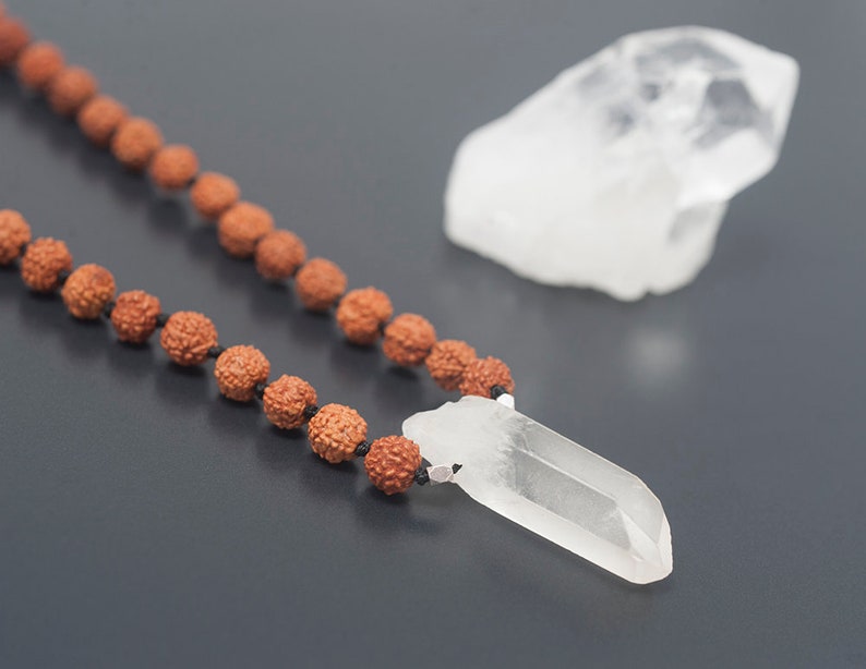 May include: A brown wooden mala bead necklace with a clear quartz crystal pendant. The necklace is lying on a dark gray surface.