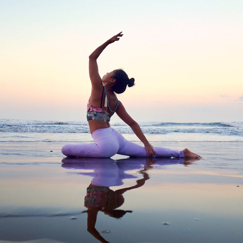 May include: A woman in a purple yoga outfit performs a split pose on a sandy beach with the ocean in the background. The sky is a soft pink and orange.
