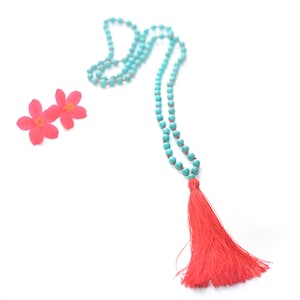 May include: A turquoise beaded necklace with a long, vibrant red tassel. Two small, bright pink flowers are positioned near the necklace. The necklace is displayed on a white background.