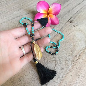 May include: A turquoise and black beaded necklace with a gold feather pendant and black tassel. The necklace is on a wooden surface.