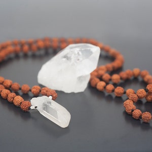 May include: A brown wooden mala bead necklace with two clear quartz crystal pendants. The necklace is laid out on a gray surface.