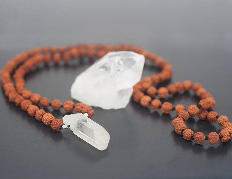 May include: A brown wooden mala bead necklace with two clear quartz crystal pendants. The necklace is laid out on a gray surface.