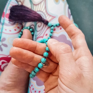 May include: A turquoise blue beaded necklace with a silver charm. The necklace is being held in a person's hands.