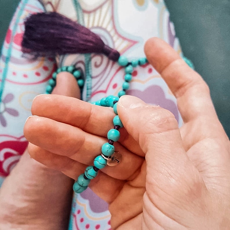May include: A turquoise blue beaded necklace with a silver charm. The necklace is being held in a person's hands.