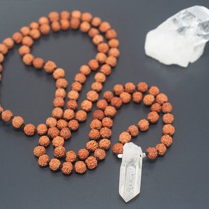 May include: A brown mala bead necklace with a clear quartz crystal pendant. The necklace is laid out on a black surface with a separate clear quartz crystal.