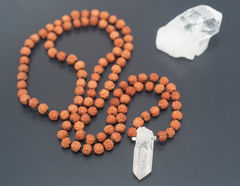 May include: A brown mala bead necklace with a clear quartz crystal pendant. The necklace is laid out on a black surface with a separate clear quartz crystal.