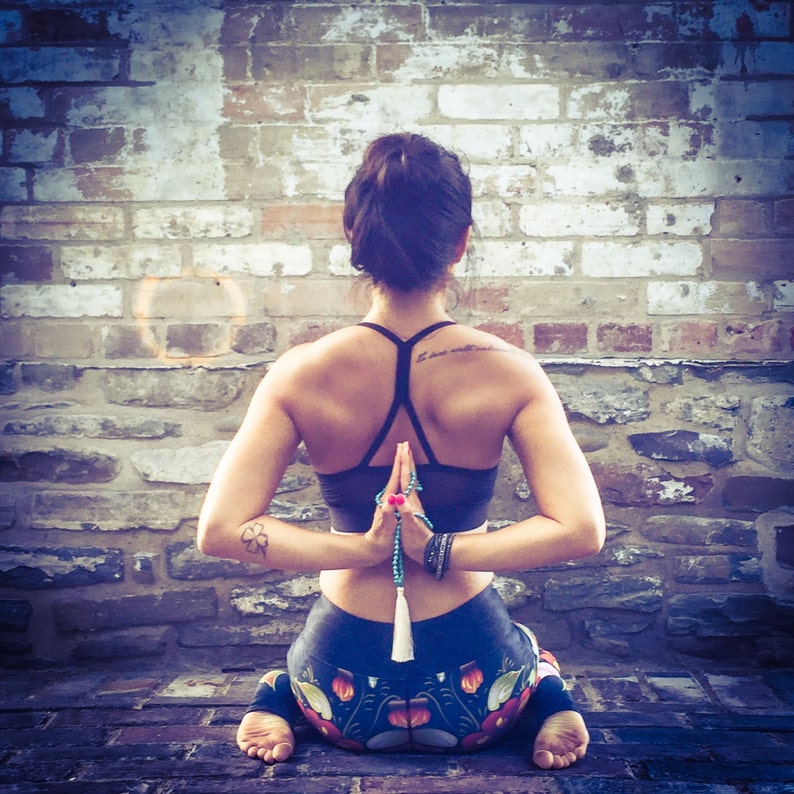 May include: A woman sits with her legs crossed and her back straight, wearing black yoga pants with a colorful floral pattern and a black sports bra. She has her hands clasped in front of her chest, with a beaded necklace hanging down from her hands. The woman is sitting in front of a brick wall.