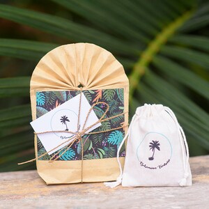 May include: A small brown paper bag with a tropical print and a white card with a palm tree logo that says "Bohemian Vibes". The bag is tied with twine and is sitting on a wooden surface. Next to the bag is a small white drawstring bag with the same palm tree logo and the text "Bohemian Vibes".
