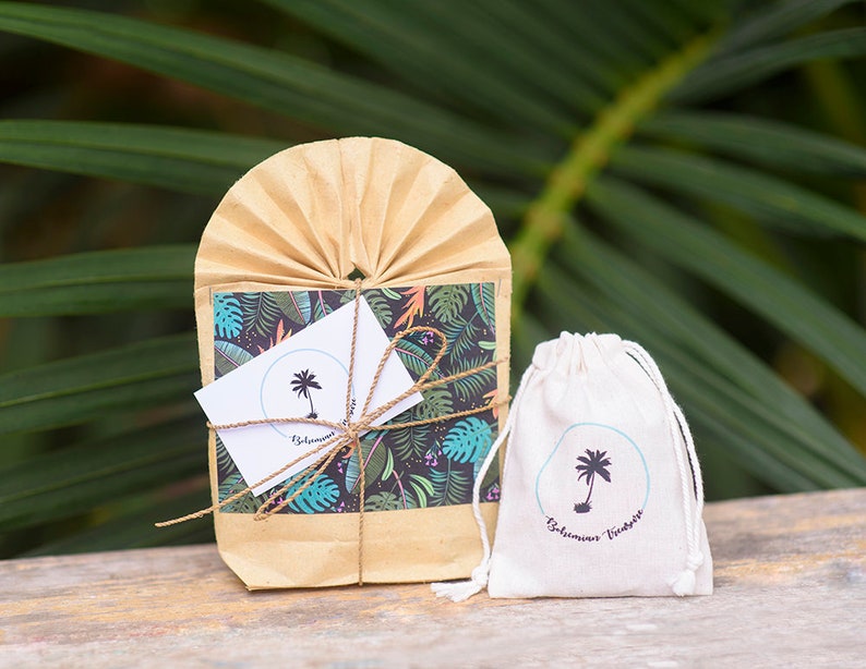 May include: A small brown paper bag with a tropical print and a white card with a palm tree logo that says "Bohemian Vibes". The bag is tied with twine and is sitting on a wooden surface. Next to the bag is a small white drawstring bag with the same palm tree logo and the text "Bohemian Vibes".