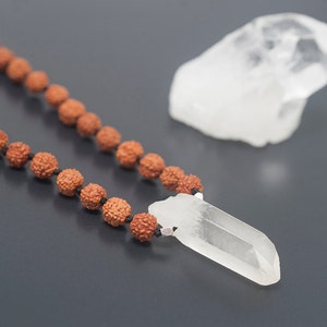 May include: A clear quartz crystal pendant on a brown wooden bead necklace. The pendant is a long, thin point with a smooth surface. The necklace is made of small, round beads that are strung together with black thread.