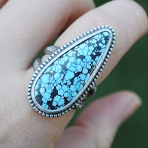 Op de afbeelding: Een zilveren ring met een grote turquoise steen in een traanvormige setting. De steen heeft een zwart en blauw marmerpatroon.