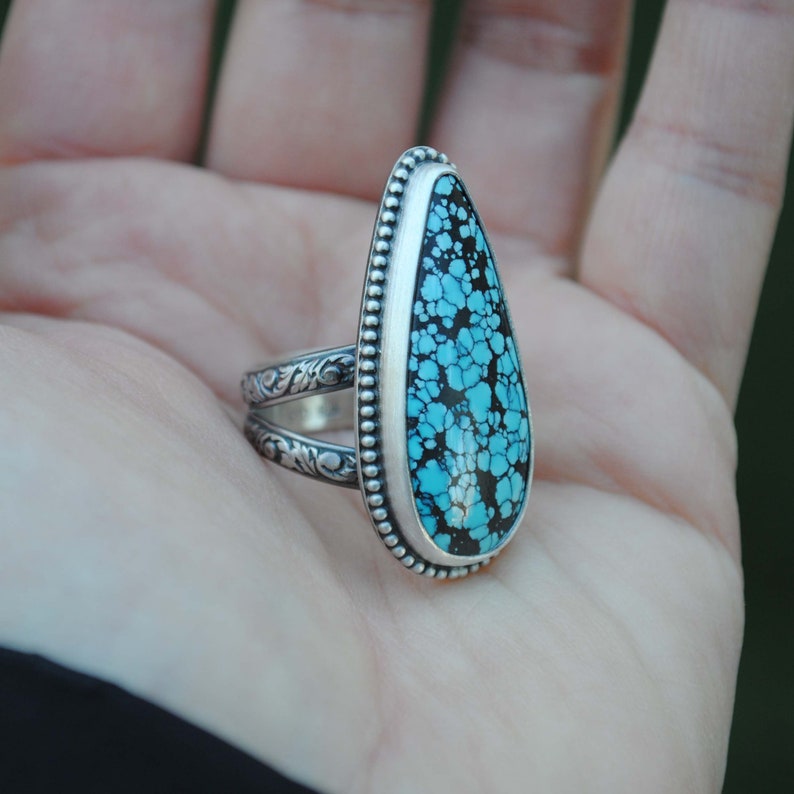 Op de afbeelding: Een zilveren ring met een traanvormige turquoise steen in een bezel setting. De steen heeft een blauw en zwart marmerpatroon.