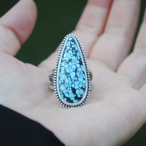 Op de afbeelding: Een zilveren ring met een traanvormige turquoise steen gezet in een geperlde bezel. De turquoise is een levendige blauwe kleur met zwarte aderen.