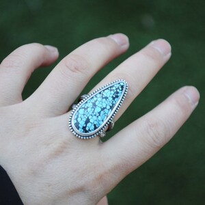 Op de afbeelding: Een zilveren ring met een traanvormige turquoise steen gezet in een zilveren bezel. De steen heeft een blauw en zwart marmerpatroon.