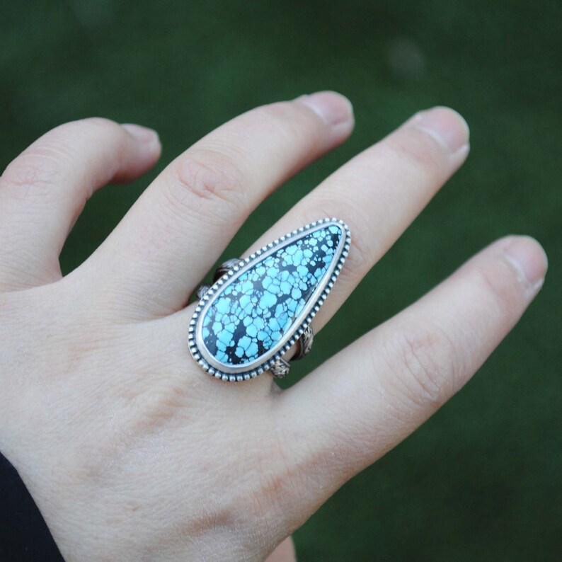 Op de afbeelding: Een zilveren ring met een traanvormige turquoise steen gezet in een zilveren bezel. De steen heeft een blauw en zwart marmerpatroon.