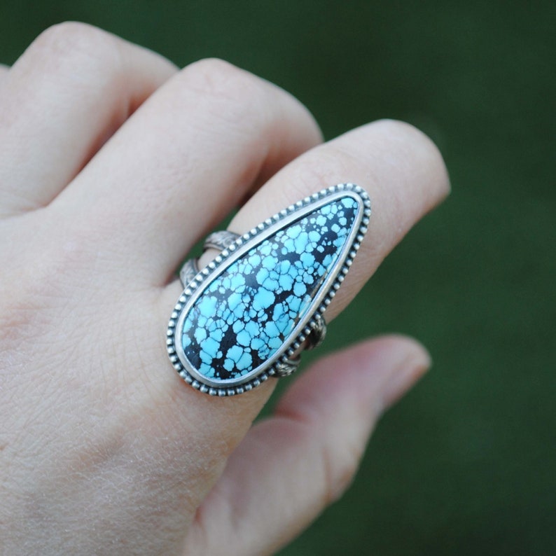 Op de afbeelding: Een zilveren ring met een grote turquoise steen in een traanvormige setting. De steen heeft een zwart en blauw marmerpatroon.