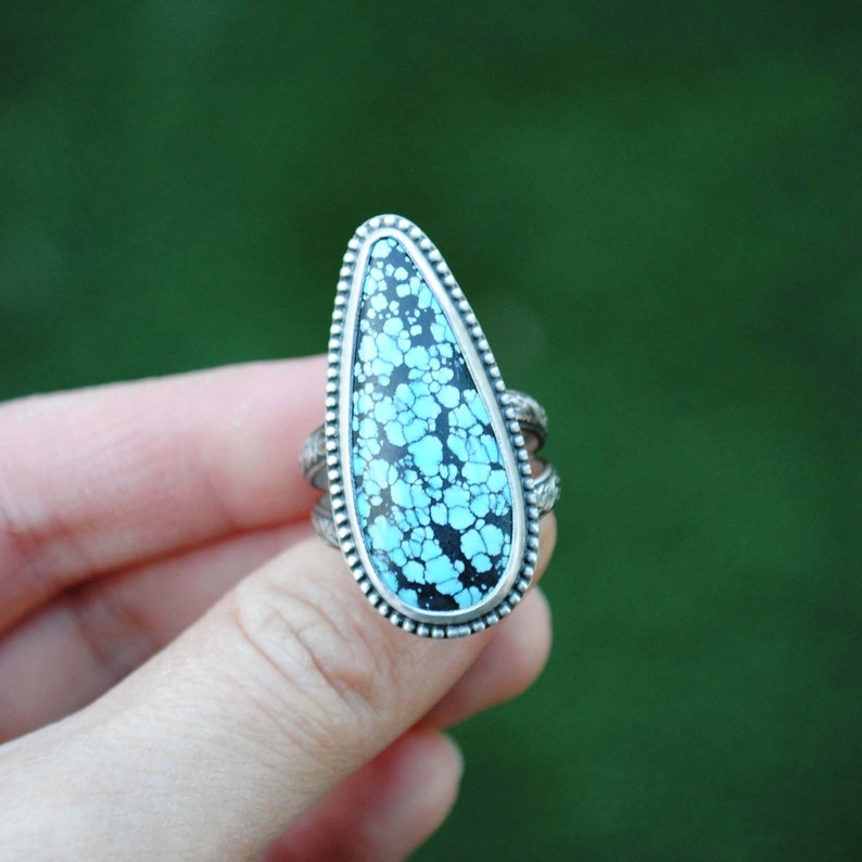 Op de afbeelding: Een zilveren ring met een traanvormige turquoise steen. De steen heeft een zwart en blauw marmerpatroon.