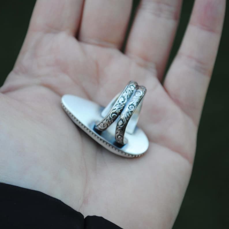 Op de afbeelding: Een zilveren ring met een brede band en een decoratief ontwerp. De ring is gemaakt van twee banden die aan de bovenkant aan elkaar zijn verbonden. De band is ovaalvormig en heeft een parelrand.