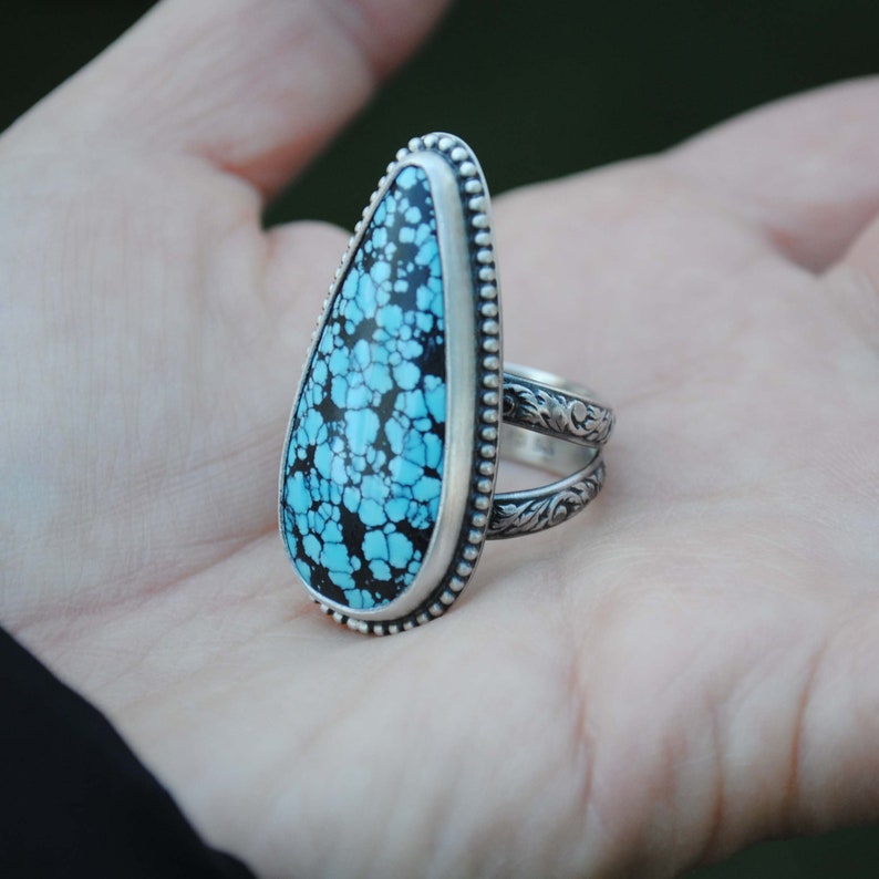 Op de afbeelding: Een zilveren ring met een traanvormige turquoise steen gezet in een geperlde bezel. De ringband is ook van zilver en heeft een ingewikkeld bloemmotief.