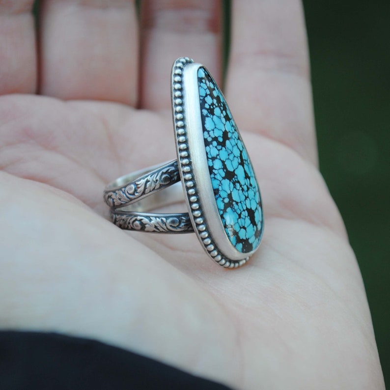 Op de afbeelding: Een zilveren ring met een turquoise steen in een traanvorm. De ring heeft een gedetailleerde band met een bloemmotief.
