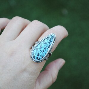 Op de afbeelding: Een zilveren ring met een grote, traanvormige turquoise steen. De steen heeft een blauw en zwart patroon.