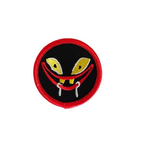 Puede incluir: Un parche bordado rojo y negro con una cara de dibujos animados con una amplia sonrisa de dientes y ojos amarillos.