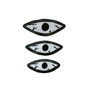 May include: Three black and white embroidered eye patches with a white background and black stitching. The eyes are outlined in black and have a black pupil.